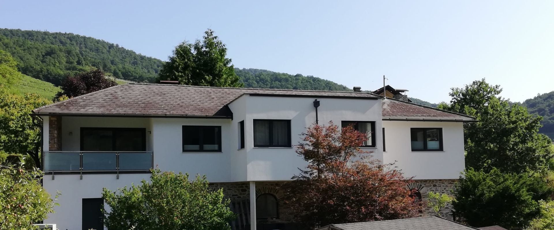 Exteriérový pohľad na moderný biely rekreačný dom so stromami v popredí a zalesnenými kopcami v pozadí.