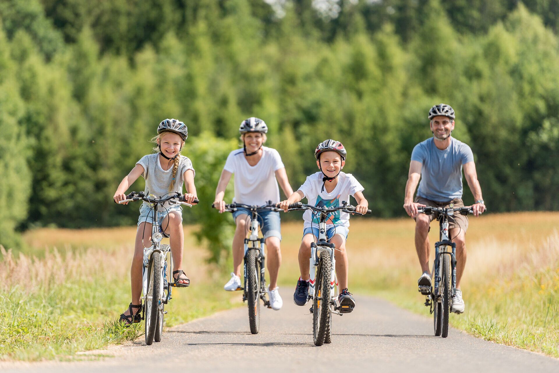Veselá rodina sa spoločne bicykluje po malebnej ceste obklopenej sviežou zeleňou a kopcami. Slnko svieti na modrej oblohe a vytvára príťažlivú atmosféru pre nezabudnuteľné zážitky v prírode.