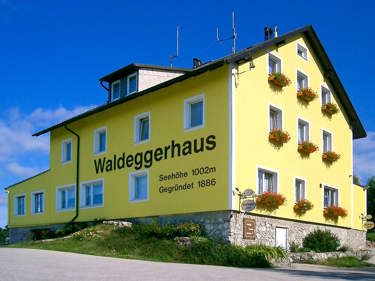 Žltá budova s nápisom "Waldeggerhaus", obklopená kvetmi.