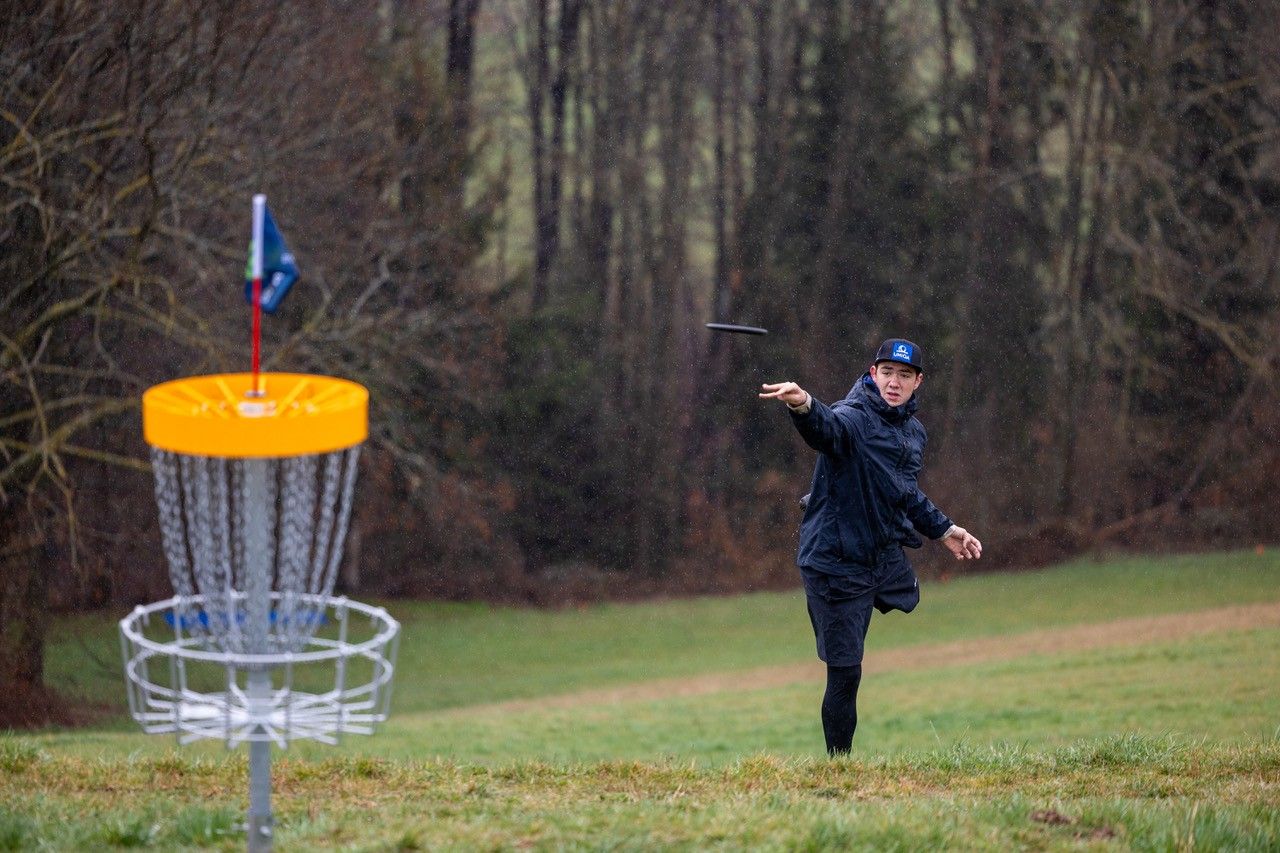 Muž hádže disk do discgolfového koša na lúke obklopenej stromami.