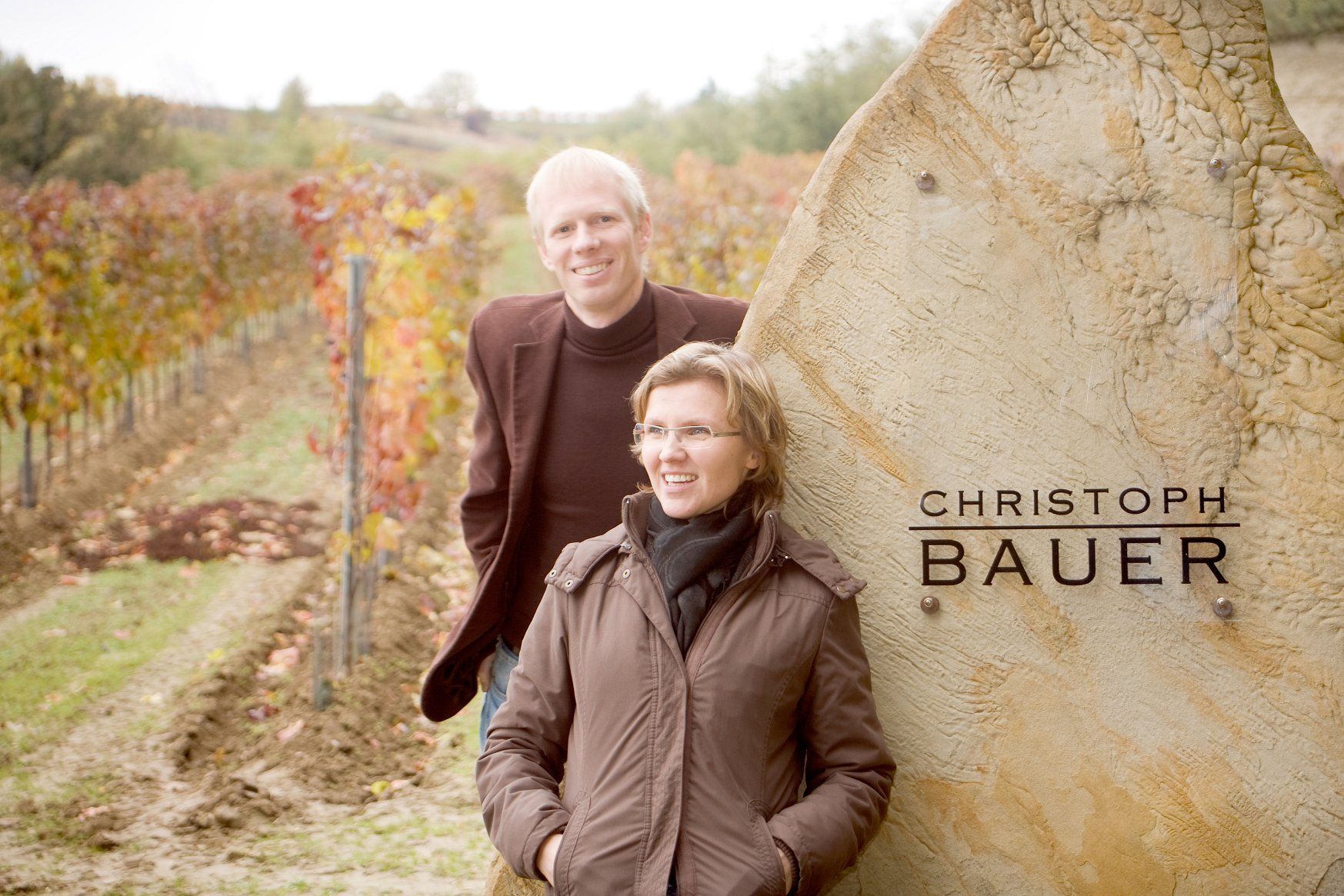 Dvaja ľudia stoja s úsmevom pred vinicou pri kameni s nápisom "Christoph Bauer".