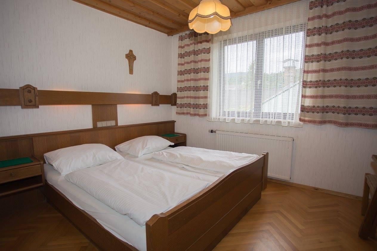 Tradične zariadená dvojlôžková izba s dreveným nábytkom, manželskou posteľou, krížom na stene a oknom so závesmi.