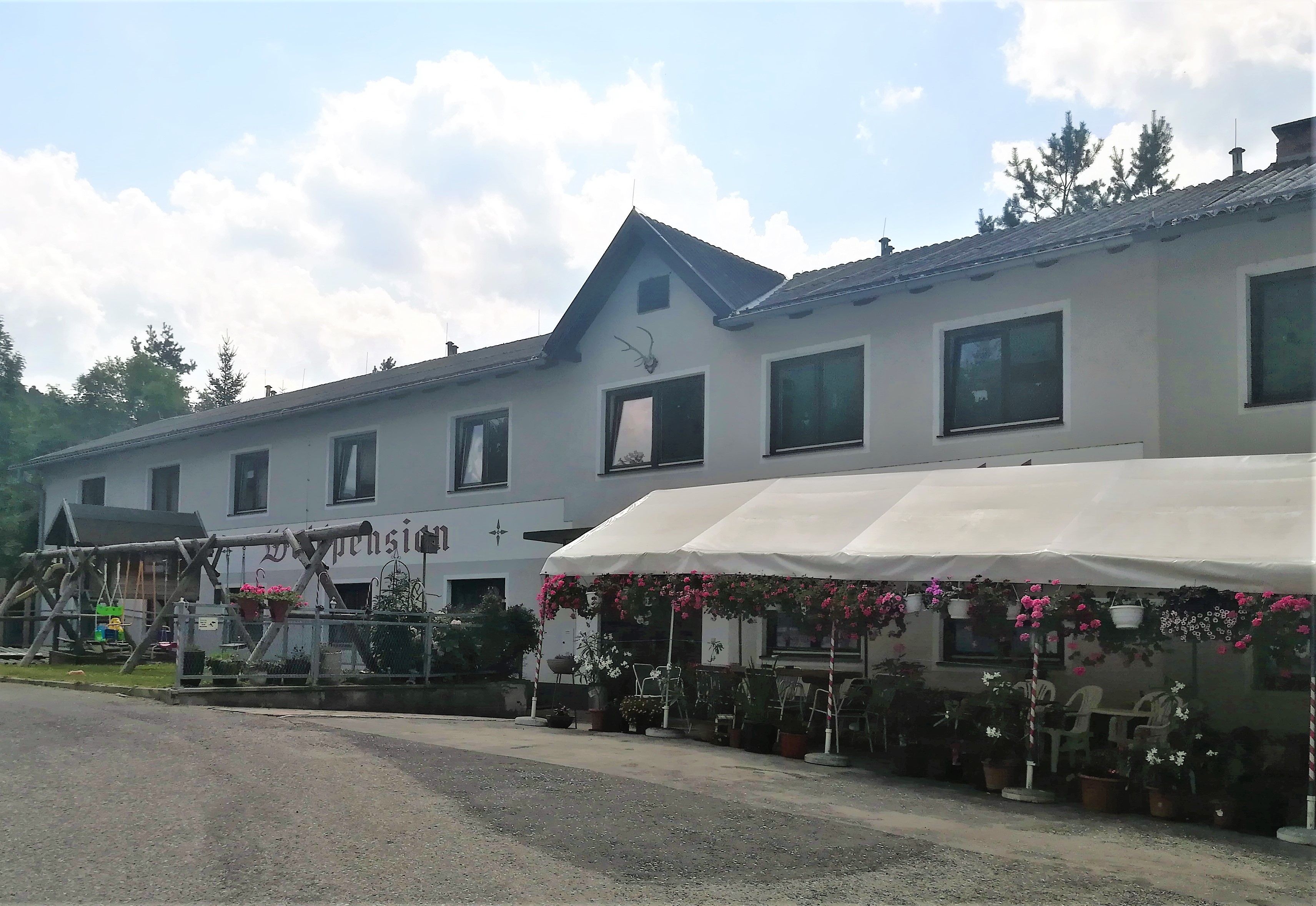 Dvojposchodová budova s nápisom "Waldpension", obklopená kvetmi a baldachýnom.