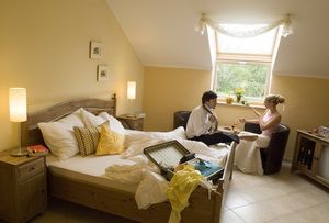 Útulná hotelová izba s párom sediacim pri stole. Posteľ je neustlaná a na posteli leží otvorený kufor.