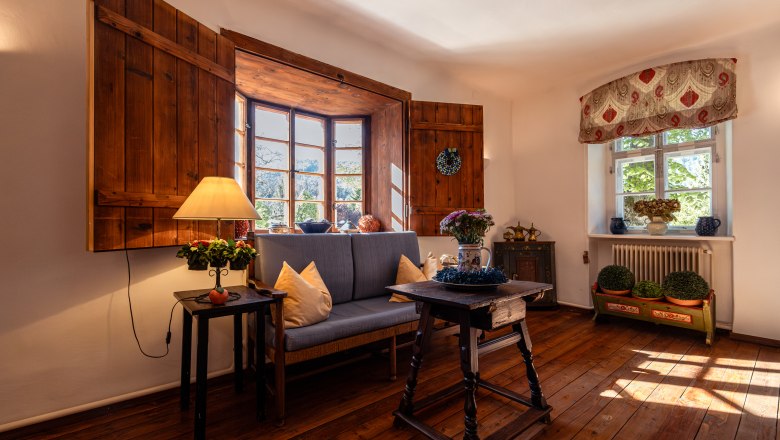 Vidiecke sídlo Oberhof, © Niederösterreich Werbung / Maximilian Pawlikowsky Útulná obývacia izba s drevenou podlahou, pohovkou, stolom a lampou pred veľkým oknom s drevenými okenicami.