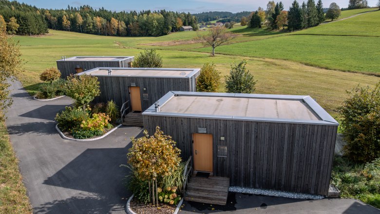 Tri moderné drevené domy v zelenej krajine so stromami a lúkami.