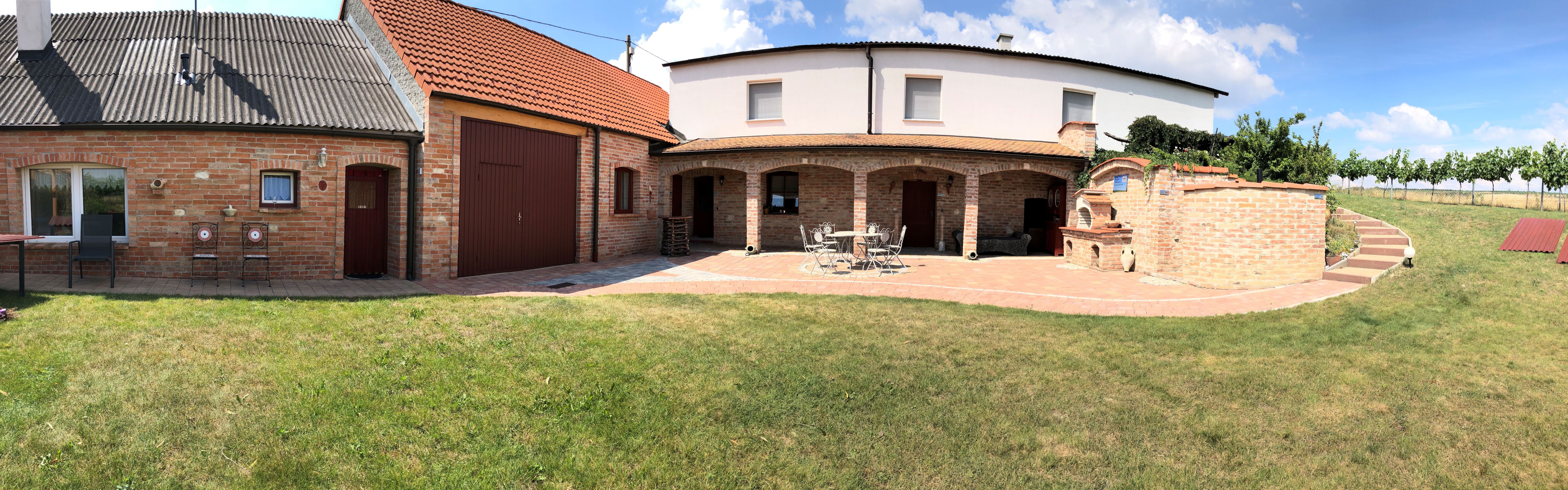 Murovaná budova so záhradou a terasou za slnečného počasia.