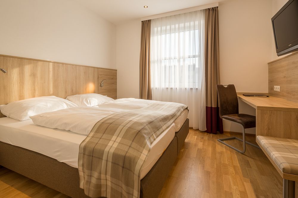 Hotelová izba s manželskou posteľou, písacím stolom a oknom so závesmi.