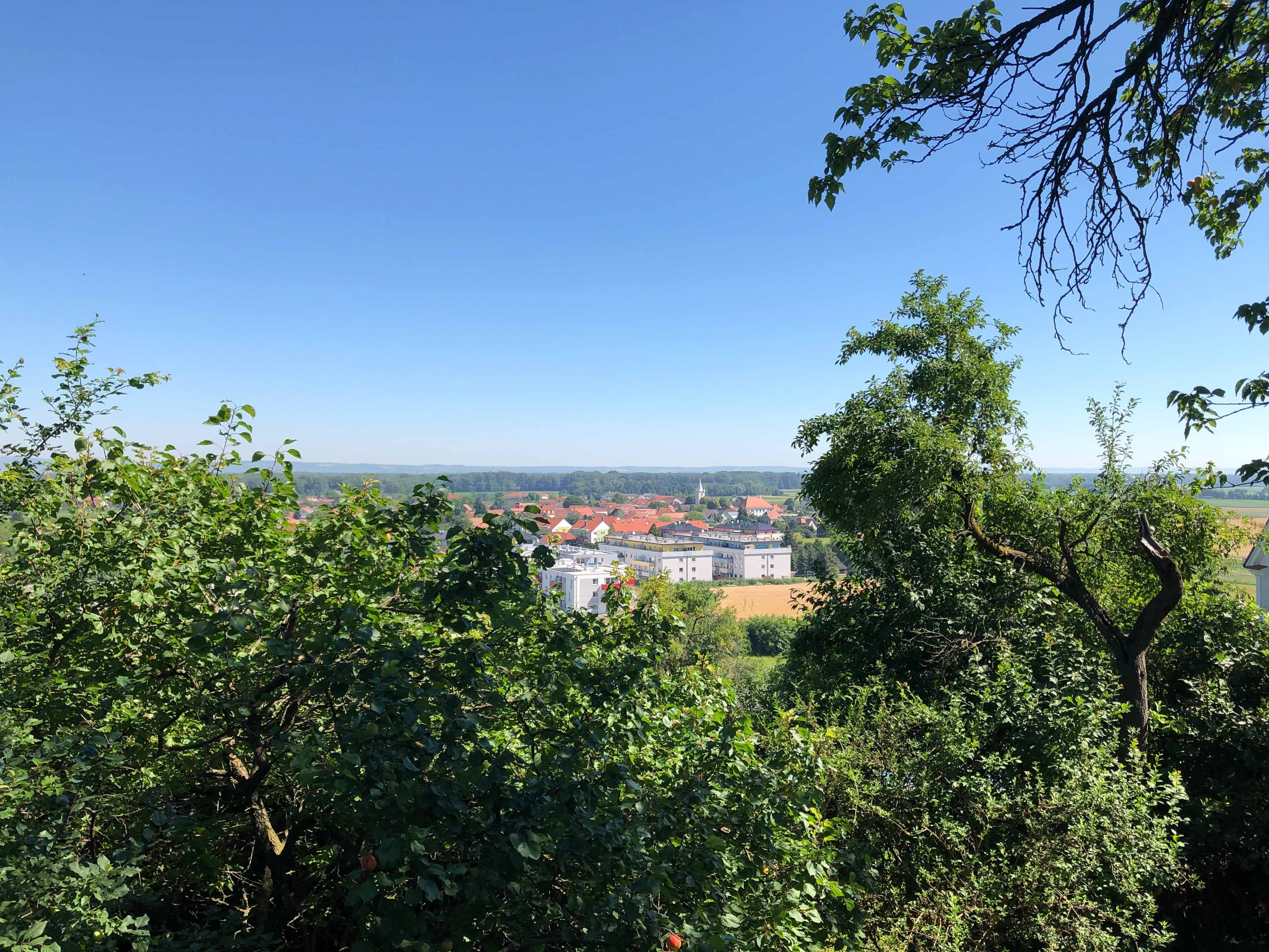 Pohľad na mesto s červenými strechami, obklopené zelenými stromami a modrou oblohou.