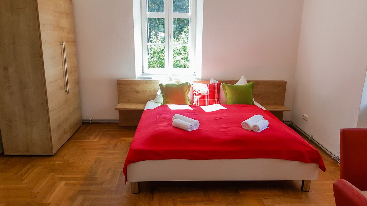Spálňa s červeným prehozom, vankúšmi a uterákmi na posteli, drevenou skriňou a oknom v pozadí.