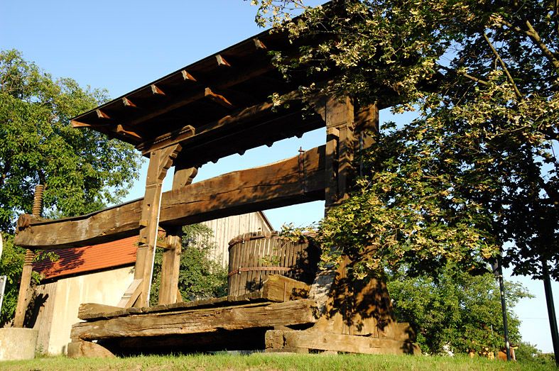 Starý drevený lis na víno vonku, obklopený stromami a budovami.