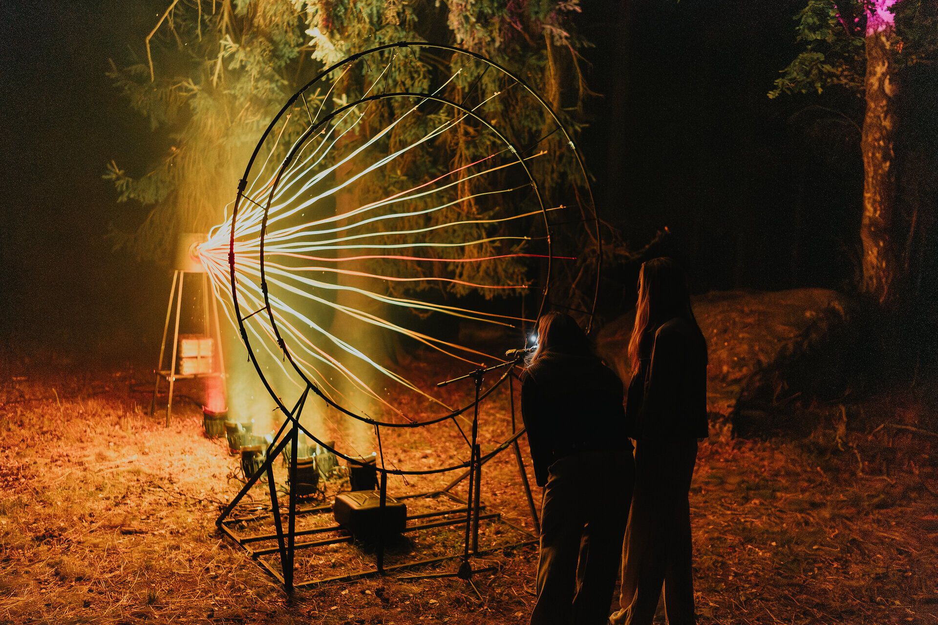 Zwei Personen stehen nachts vor einer großformatigen Lichtinstallation aus kreisförmigem Metallrahmen und farbig leuchtenden Linien; die Szene ist Teil des Lichtkunstfestivals „Blockheide leuchtet“ im Waldviertel.