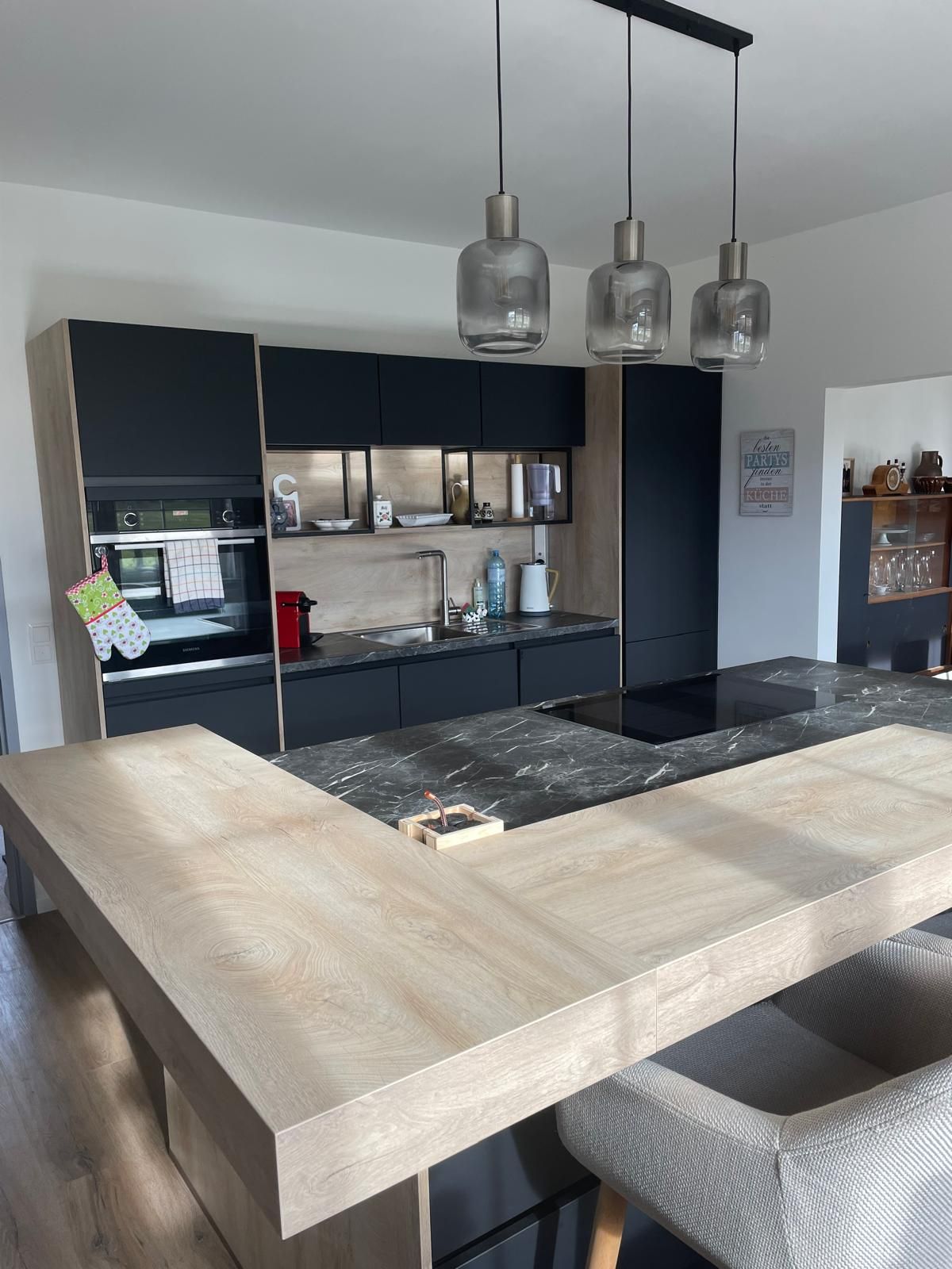 Moderná kuchyňa s drevenými a mramorovými prvkami, tromi závesnými lampami a čiernymi skrinkami.