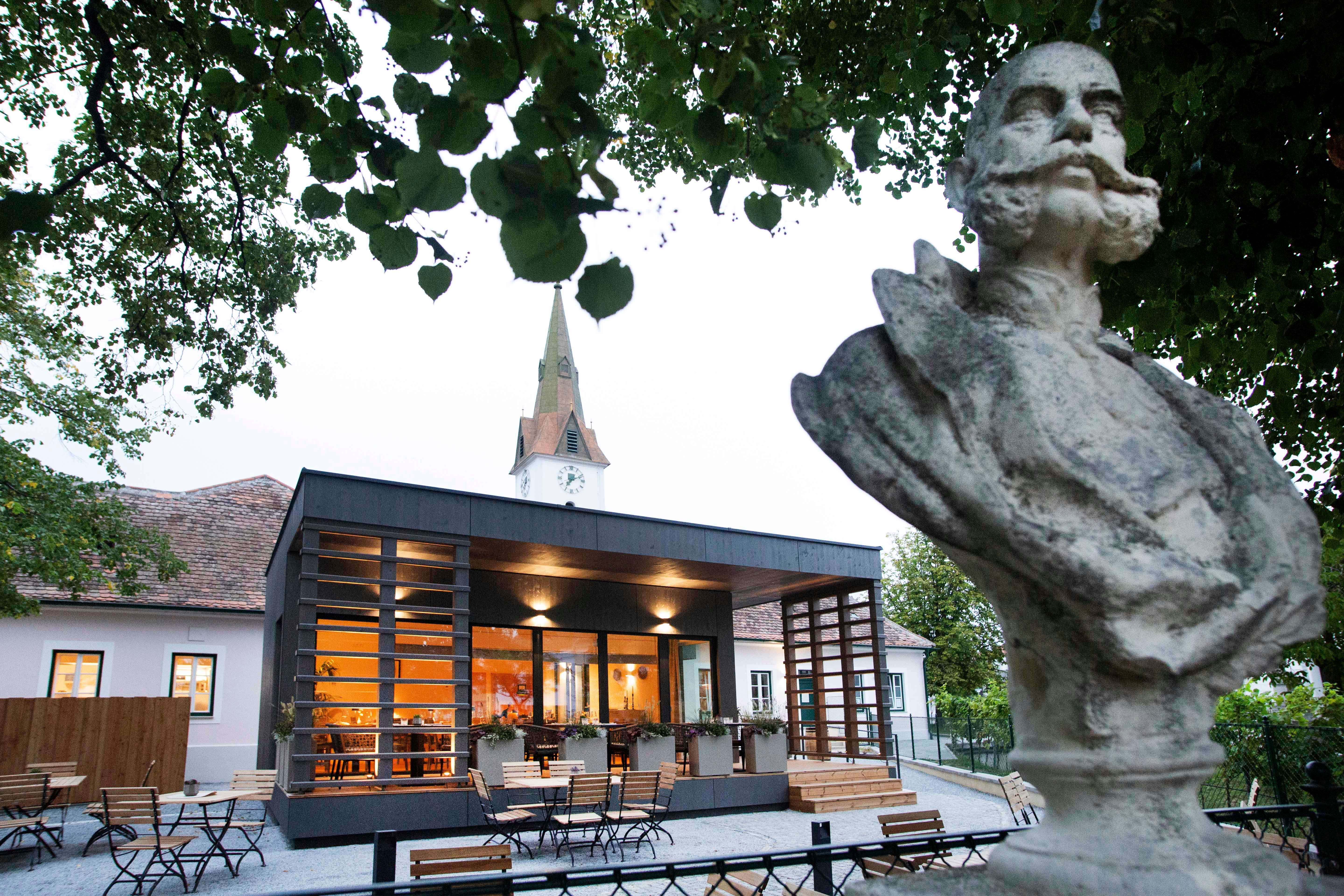 Busta pred modernou budovou s kostolnou vežou v pozadí, obklopená stromami.