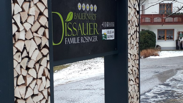 Znak farmy so stohmi dreva a domom v pozadí.