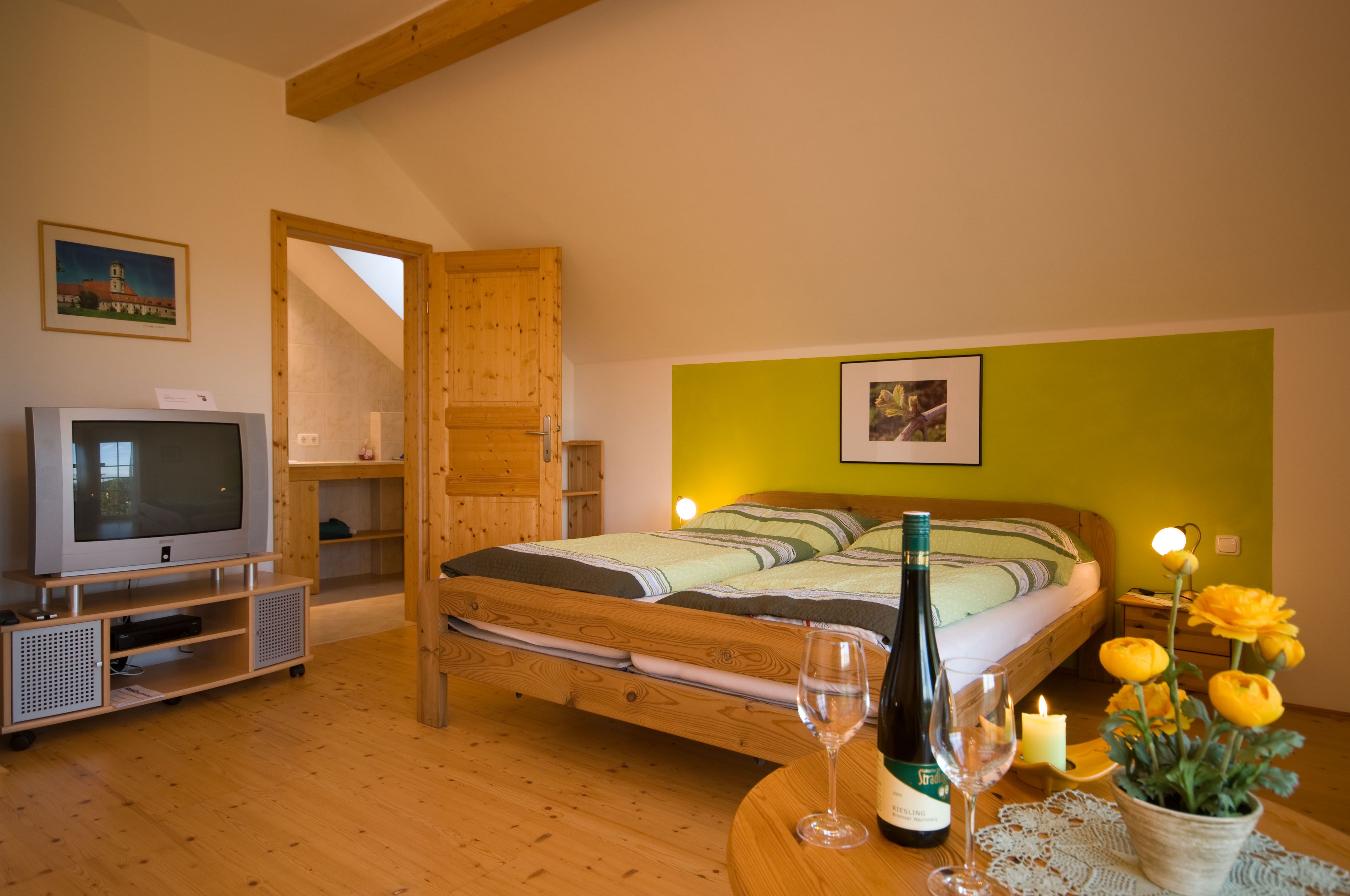 Útulná izba s manželskou posteľou, TV, fľašou vína a kvetmi na stole.