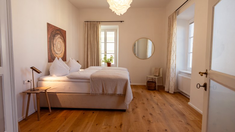 &Uacute;tuln&aacute; sp&aacute;lňa s manželskou posteľou, drevenou podlahou, umeleck&yacute;mi dielami na stene, okr&uacute;hlym zrkadlom a rastlinami na parapete.