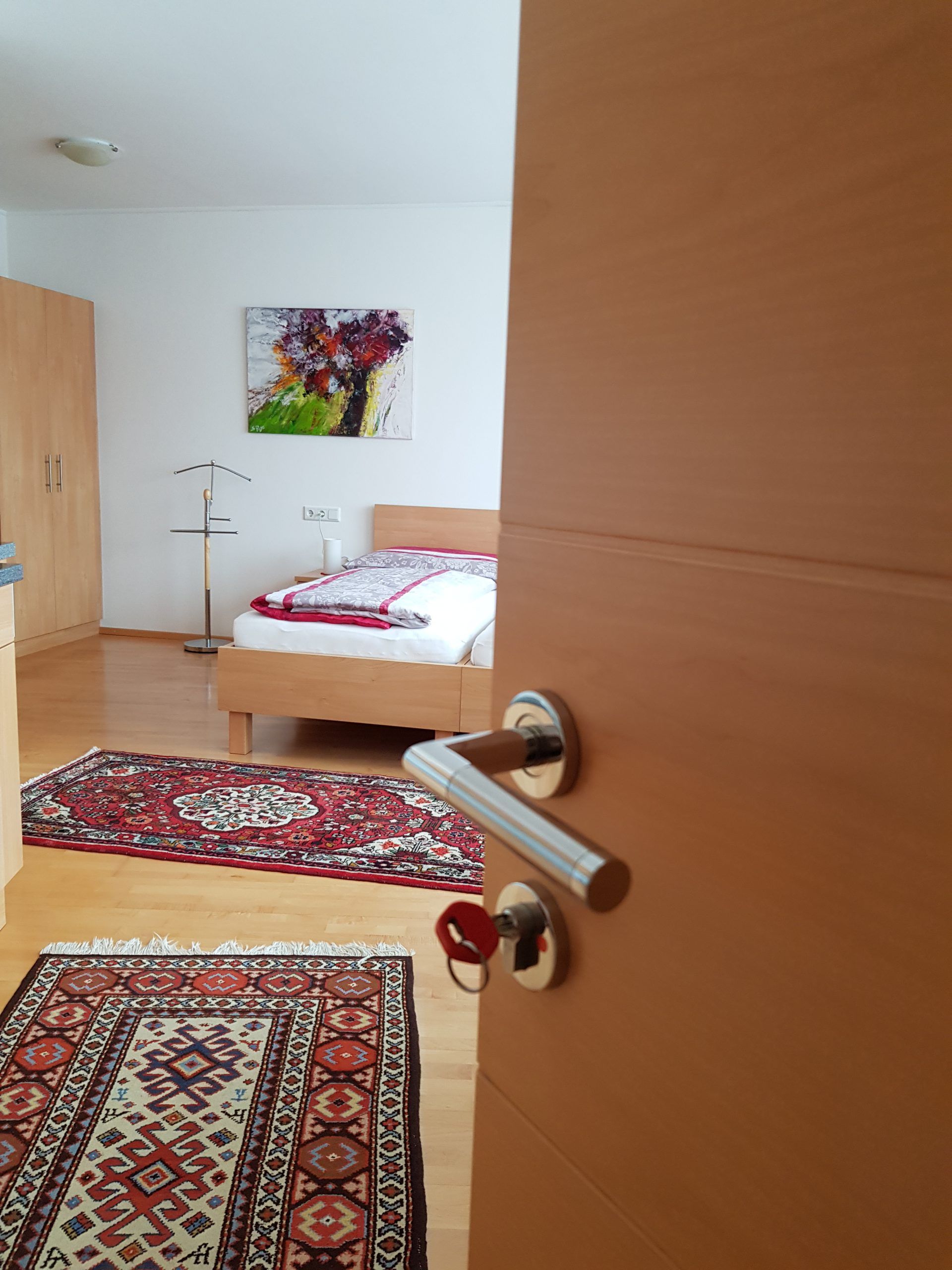 Otvorená izba s posteľou, kobercami a obrazom na stene.