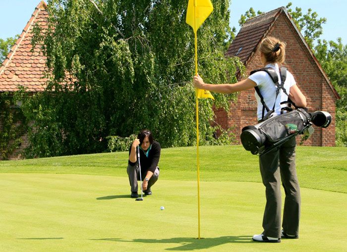 Dve ženy hrajúce golf na zelenom golfovom ihrisku, jedna drží vlajku, druhá mieri na jamku.