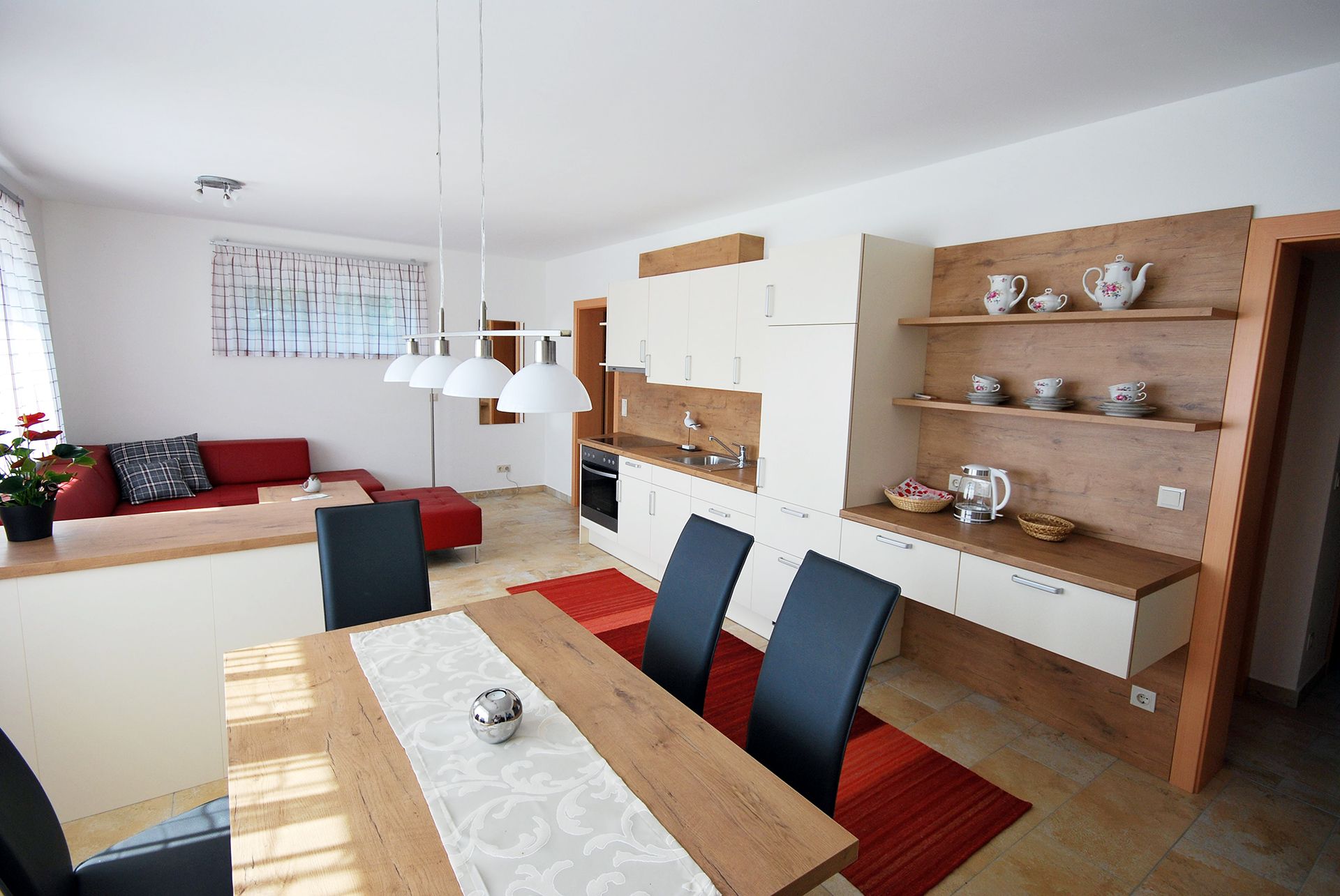 Moderný obývací a jedálenský priestor s otvorenou kuchyňou, červenou pohovkou, dreveným stolom a bielymi závesnými lampami.