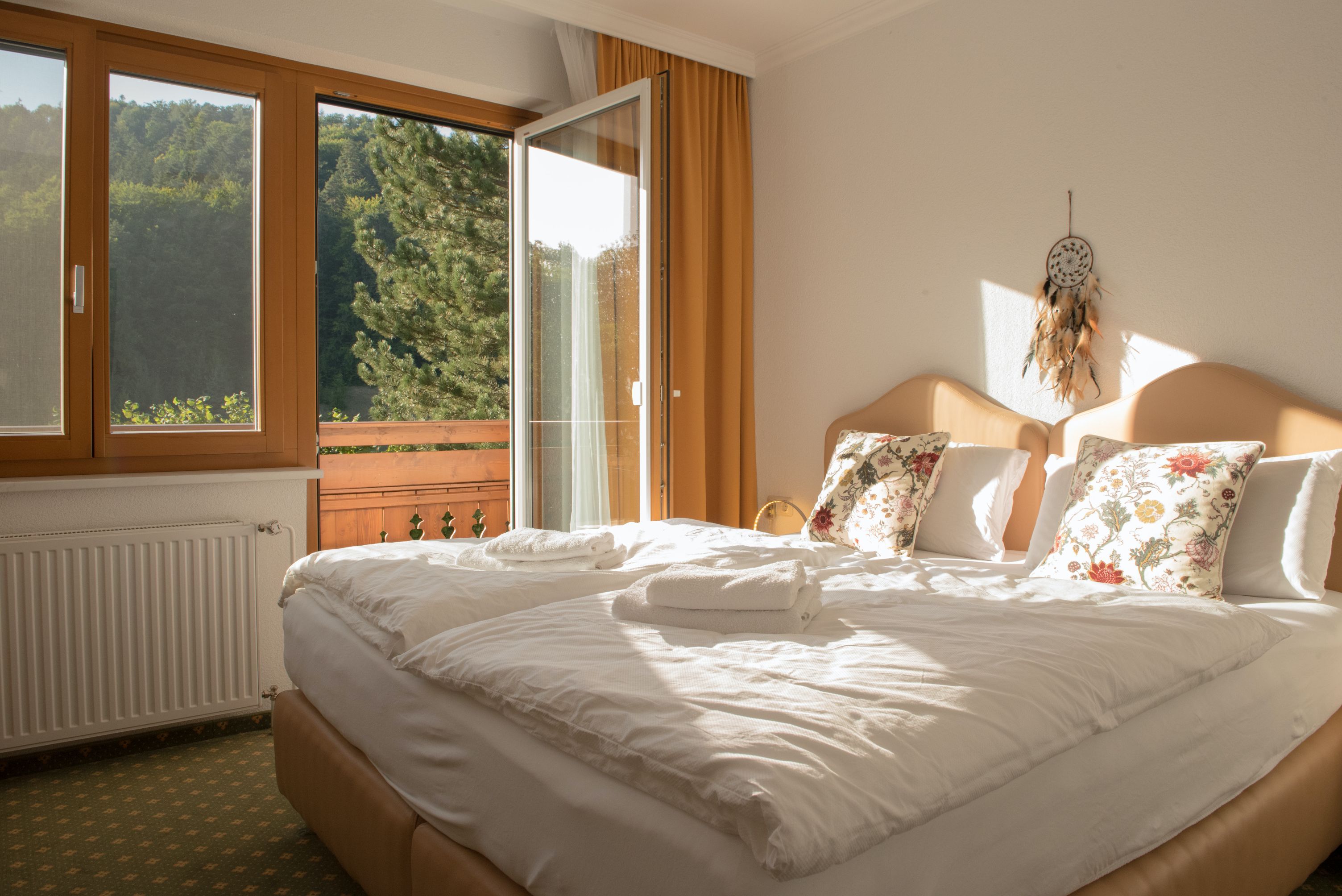 Útulná jednolôžková izba s manželskou posteľou, ozdobnými vankúšmi a otvoreným balkónom s výhľadom na zelené stromy.