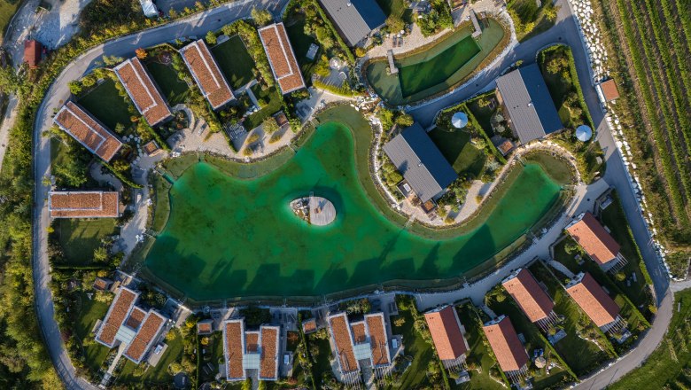 Leteck&yacute; pohľad na chaty okolo zelen&eacute;ho jazera s m&oacute;lom uprostred.