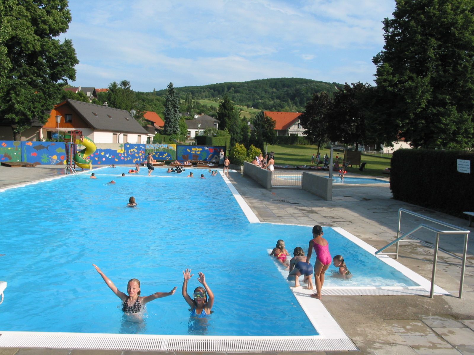 Vonkajší bazén s plavákom a šmykľavkou, obklopený stromami a lúkou.