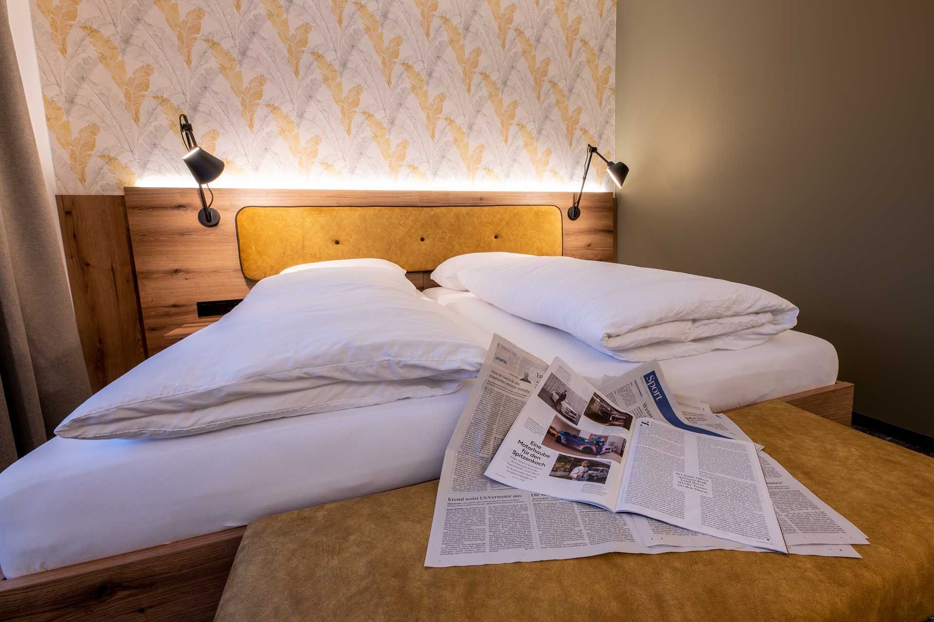 Útulná izba pre hostí s manželskou posteľou, novinami na perinách a štýlovými tapetami v pozadí.