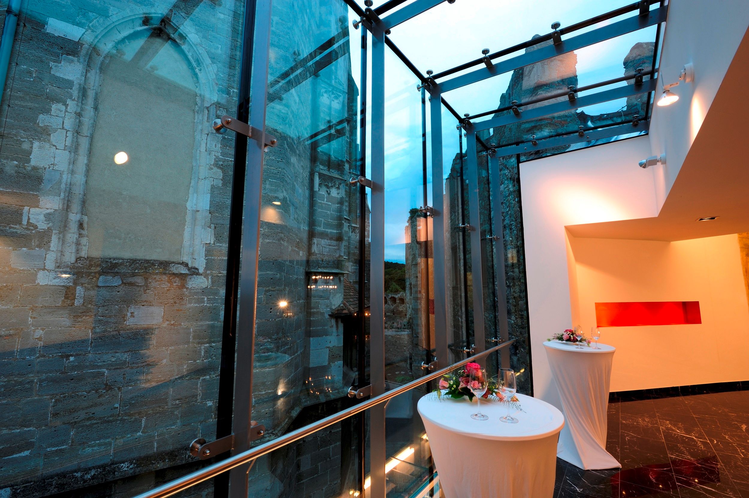 Moderná sklenená architektúra vedľa starej kamennej steny so zdobenými barovými stolmi.