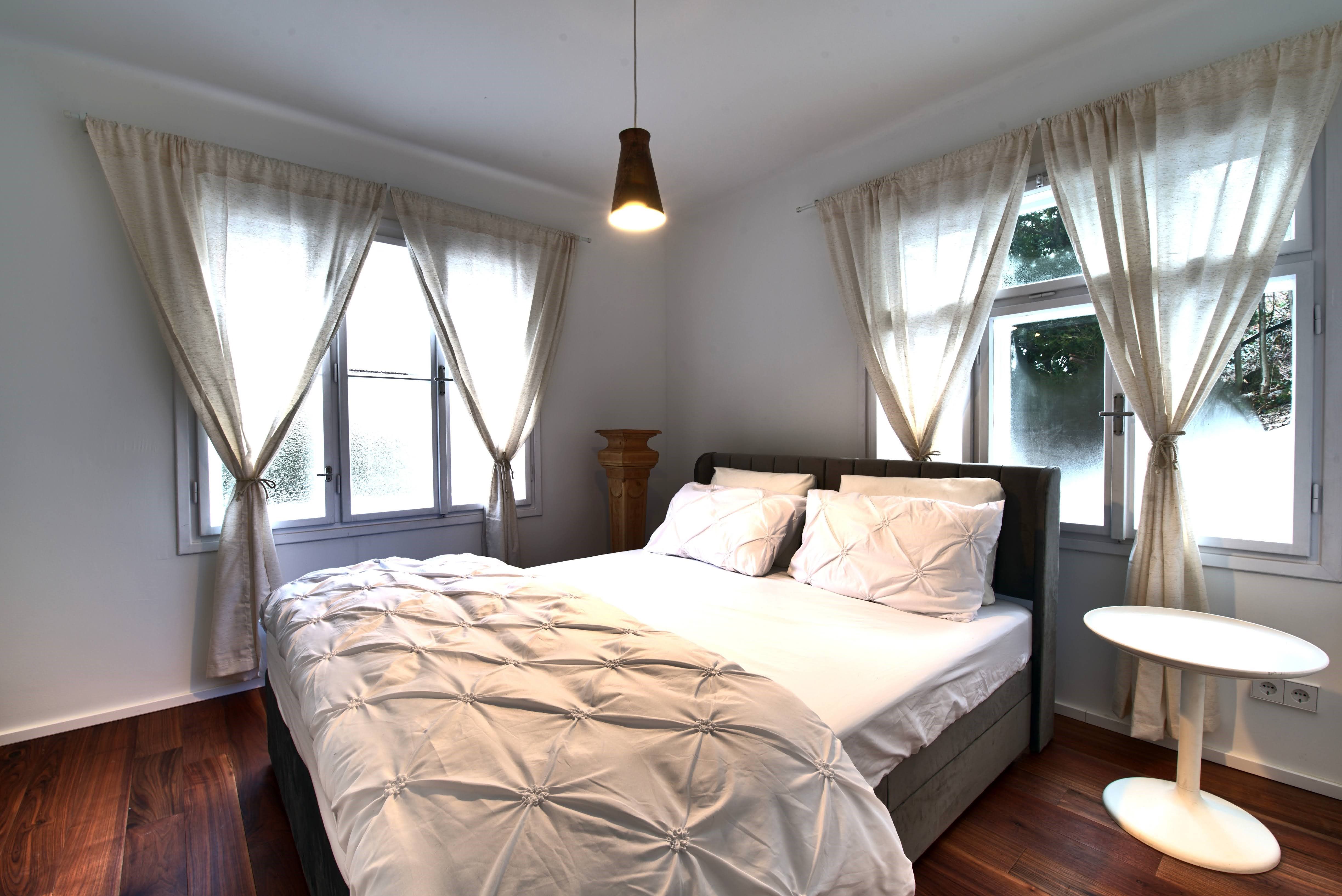 Útulná spálňa s veľkou posteľou, bielymi závesmi a malým okrúhlym stolíkom.