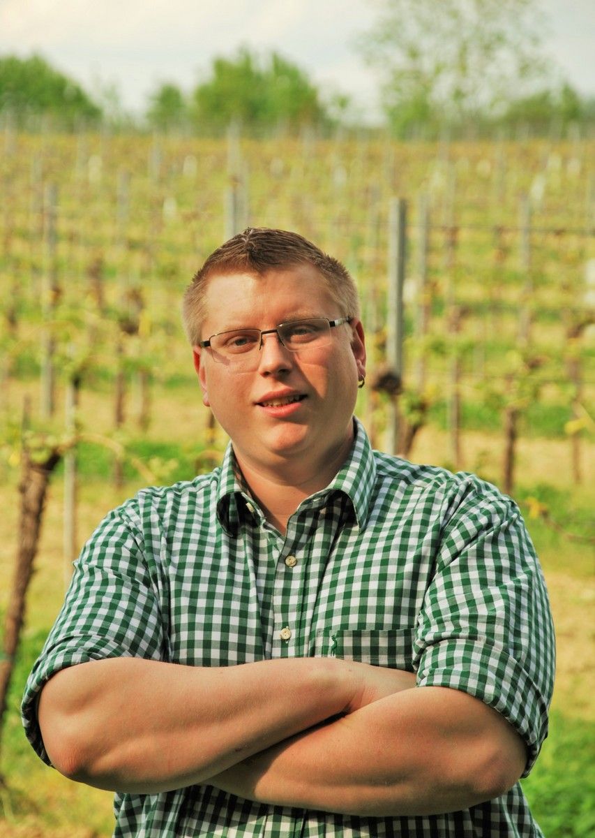 Muž v okuliaroch a košeli stojí so založenými rukami pred vinicou.