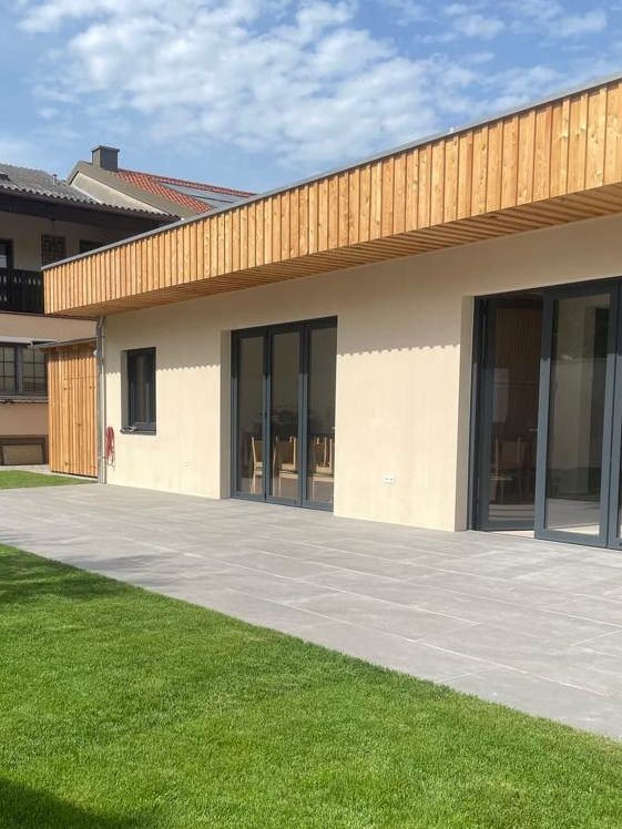 Moderná budova s dreveným obložením a veľkými oknami, obklopená terasou a trávnikom.