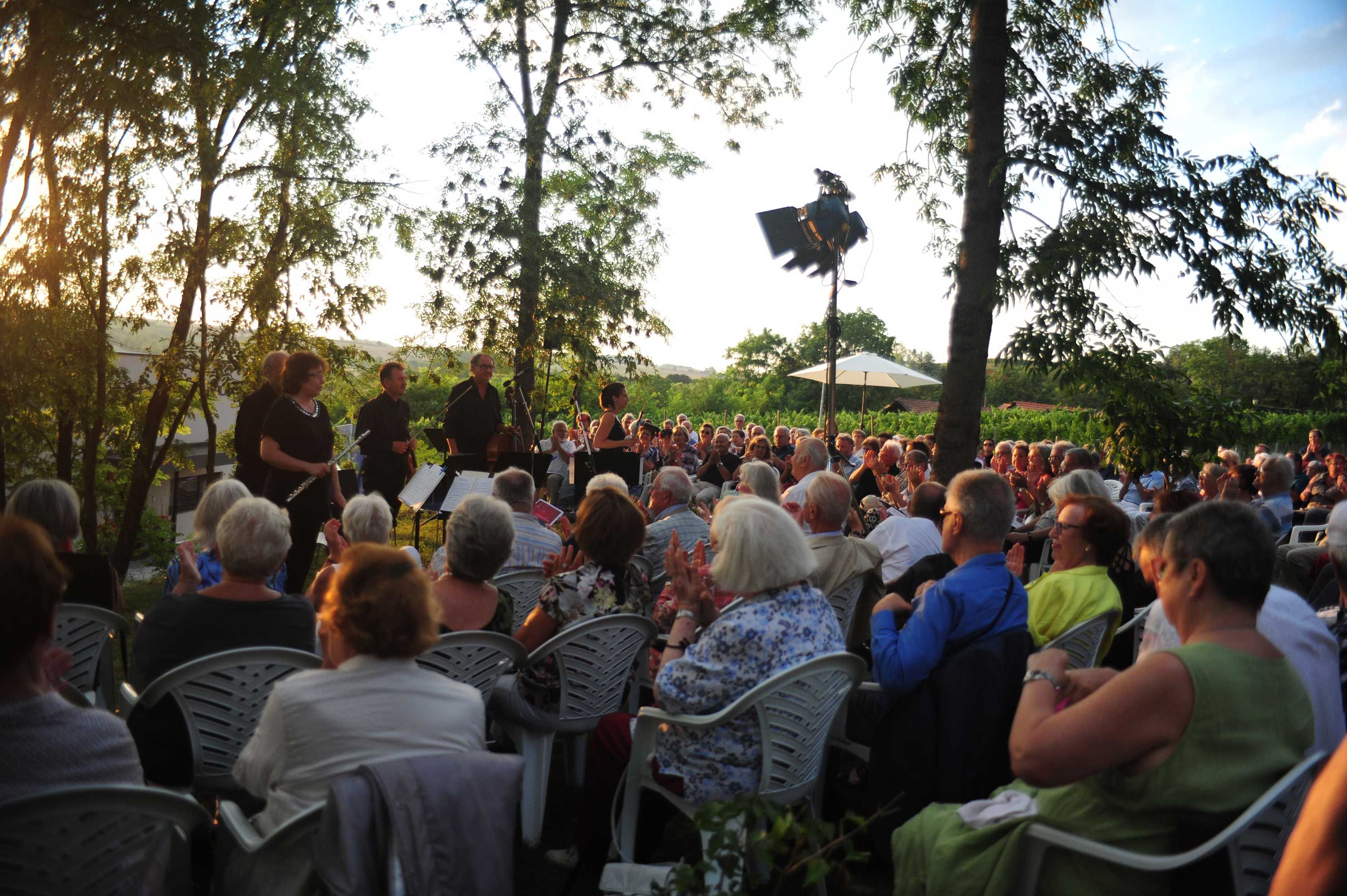 Koncert pod holým nebom s orchestrom a publikom pri západe slnka.