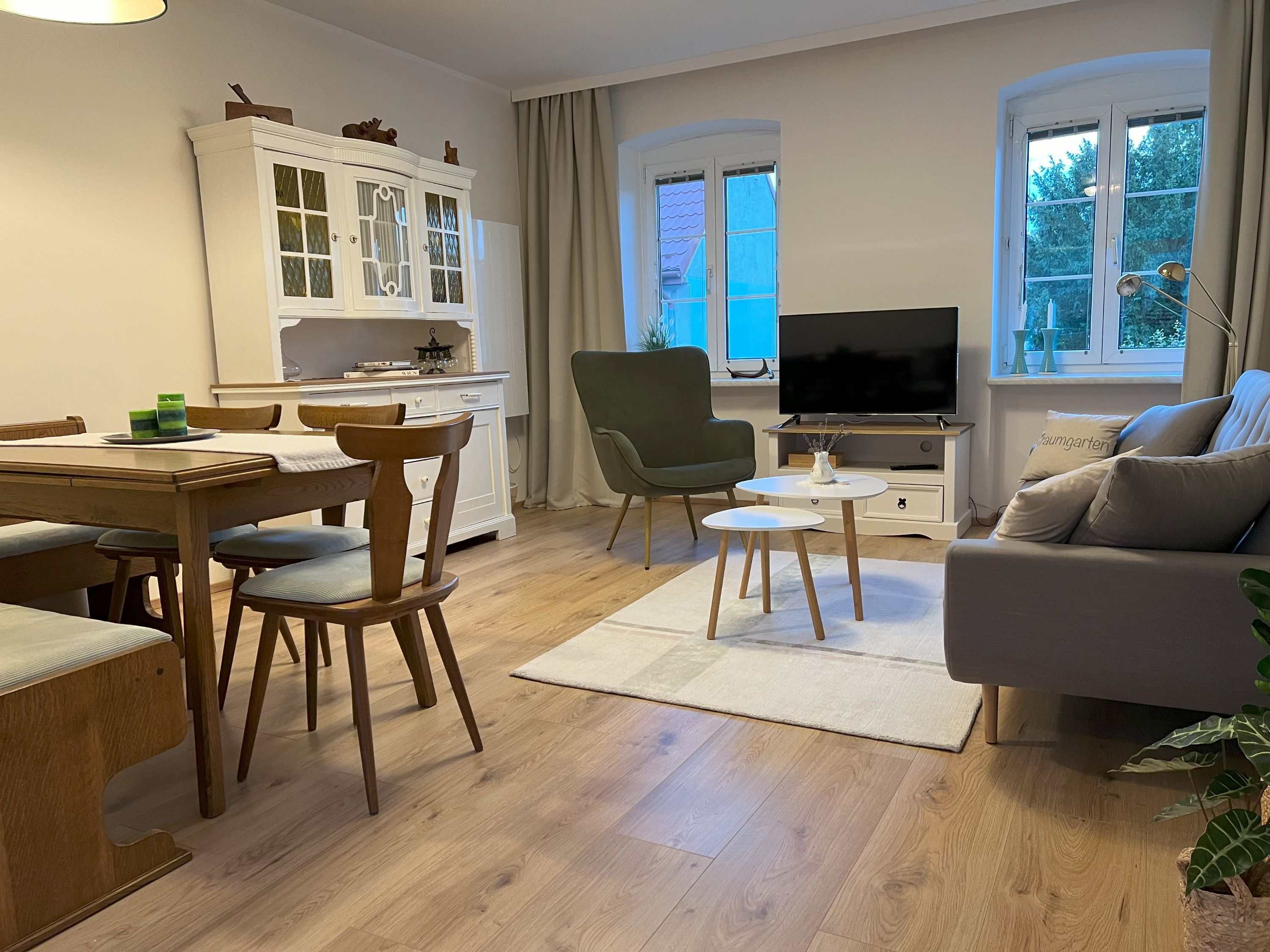 Útulná obývacia izba s dreveným nábytkom, pohovkou, kreslom, jedálenským stolom a TV. Svetlé steny a drevená podlaha.