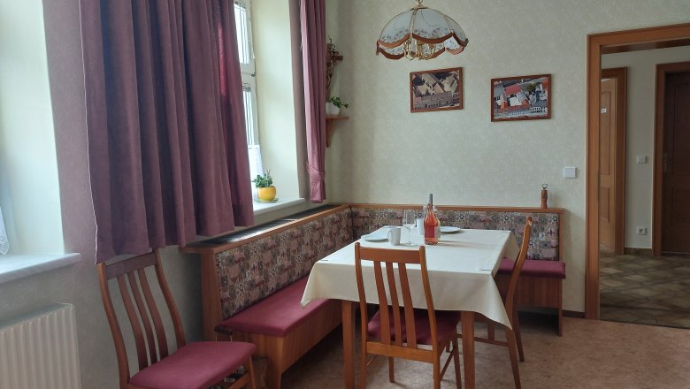 &Uacute;tuln&aacute; kuchyňa s jed&aacute;lensk&yacute;m stolom, stoličkami a rohovou lavicou. Okno so z&aacute;vesmi, obrazy na stene.