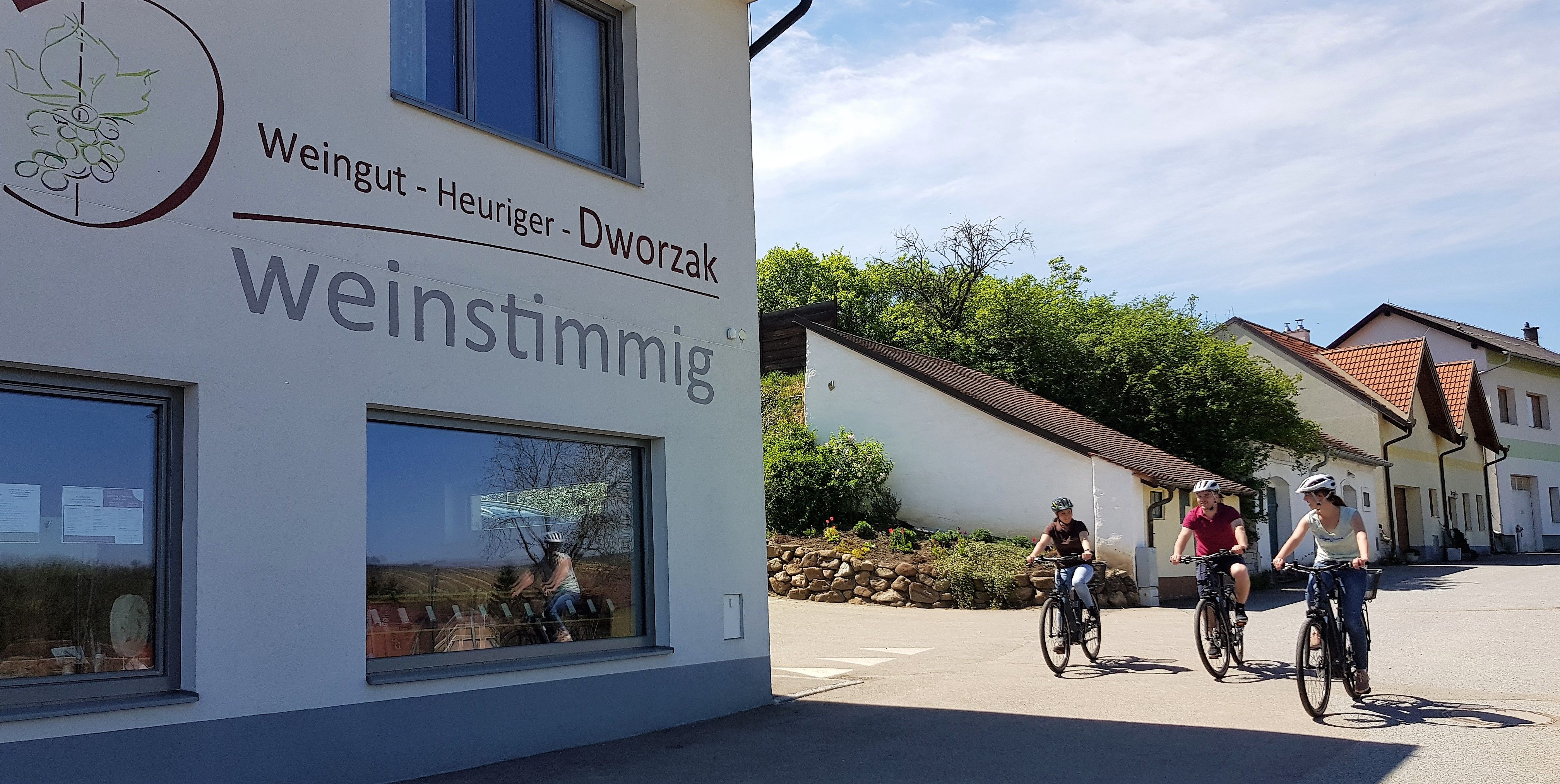 Tri osoby na bicykloch prechádzajú okolo budovy s nápisom "Weingut - Viecha - Dworzak weinstimmig".