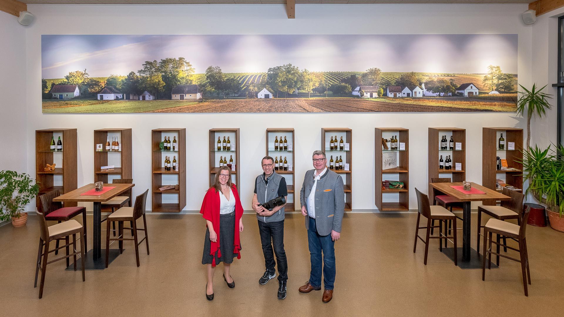 Traja ľudia stoja vo vinárni pred policami s fľašami vína a veľkou nástennou maľbou vinohradov.