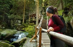 Ein sanfter Wasserfall plätschert über moosbedeckte Steine, während die umgebenden Bäume in sattem Grün erstrahlen. Die Wanderin genießt den Anblick und die frische Luft, die den Sommer in den Bergen so besonders macht.