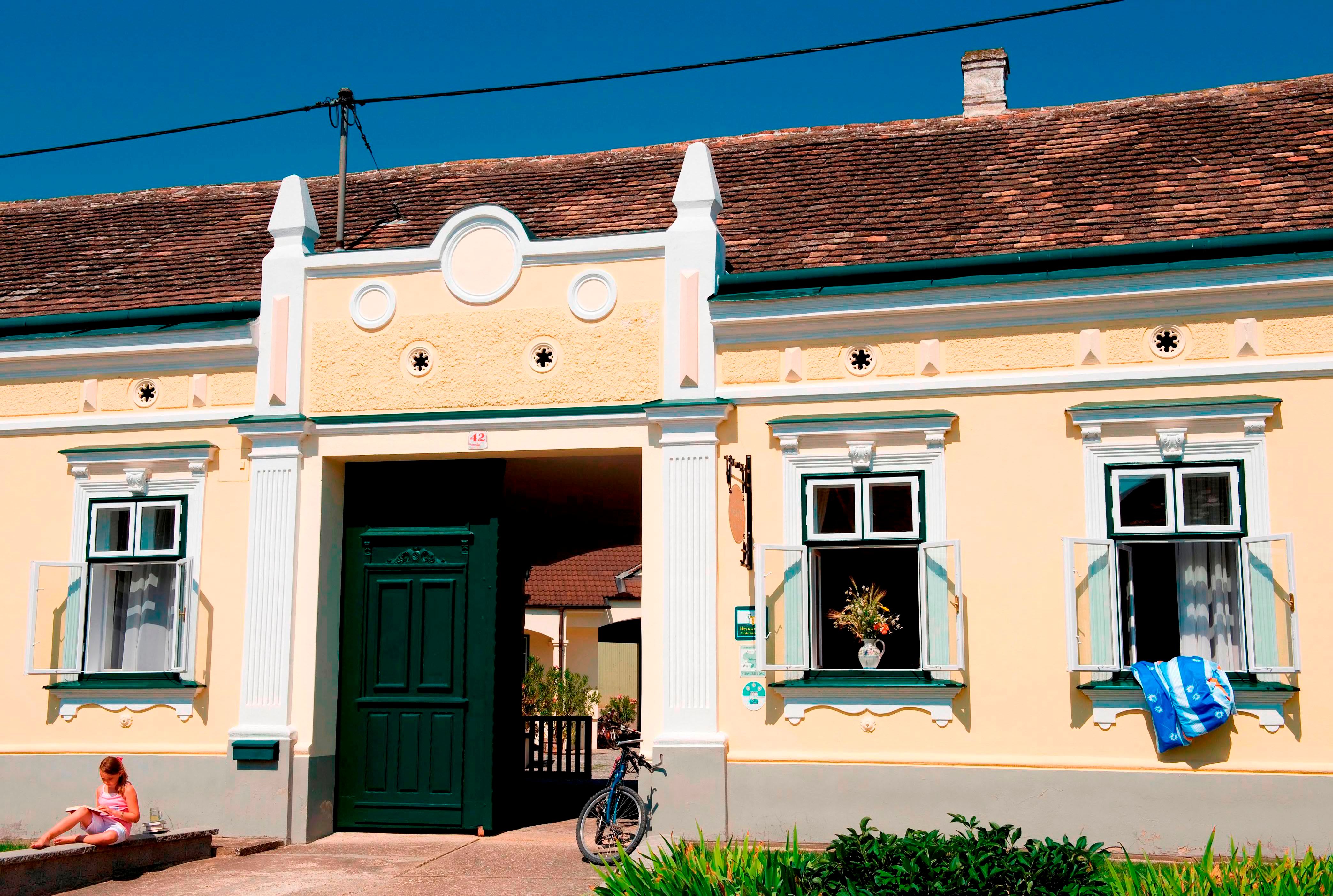Tradičná žltá budova so zelenými dverami a oknami, pred ňou bicykel a na schodoch sedí dieťa.