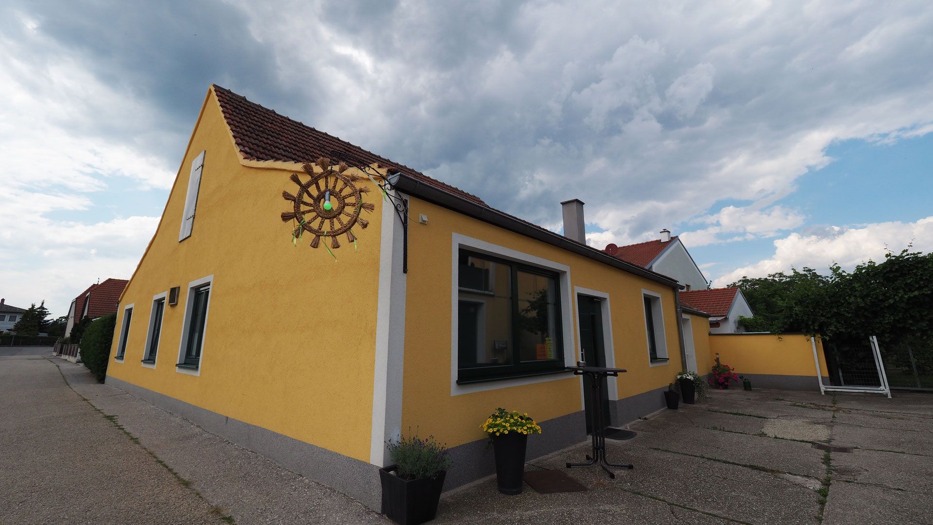 Žltá budova s červenými škridlami a ozdobným kolesom na stene, obklopená rastlinami a zamračenou oblohou.