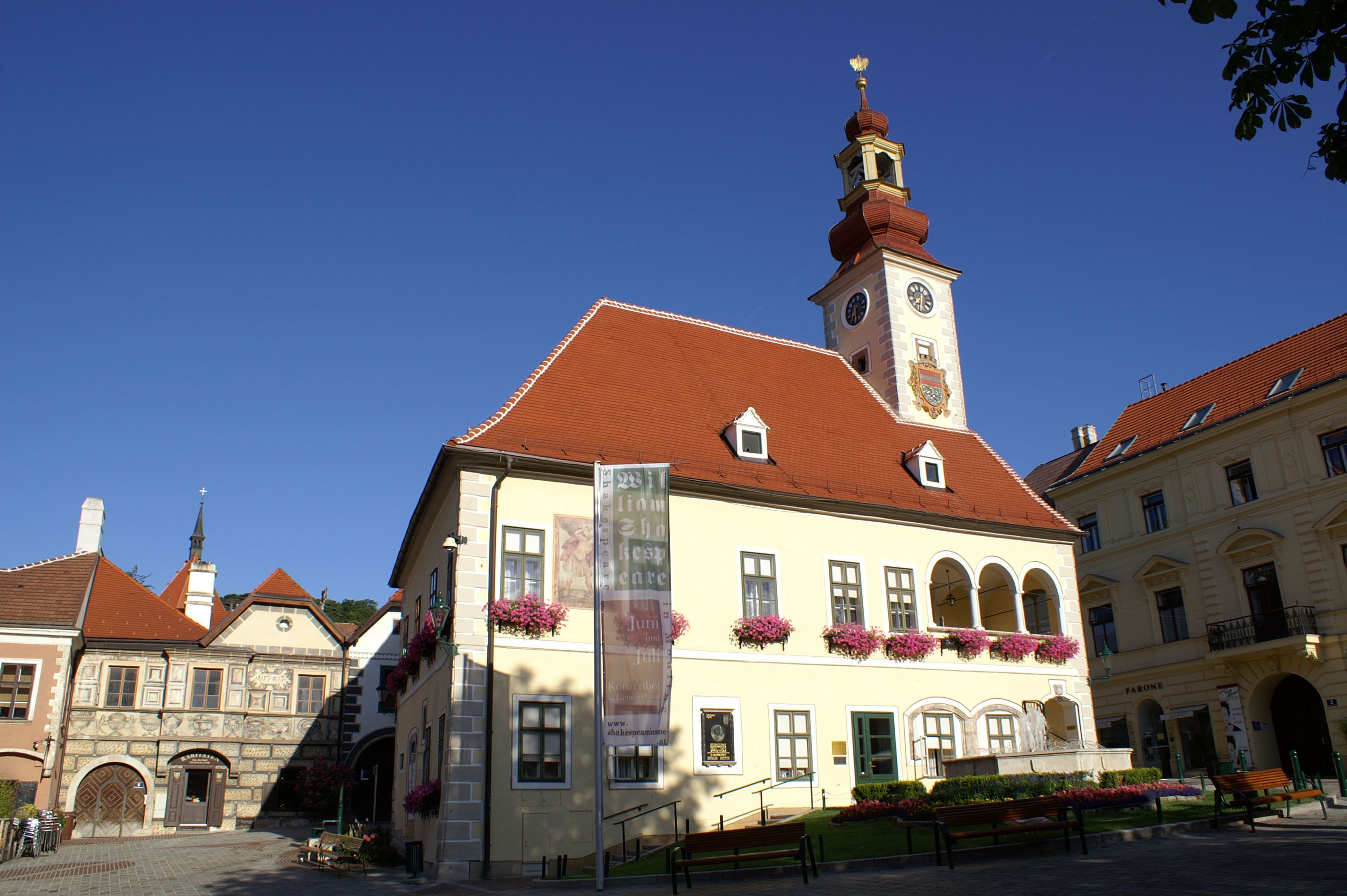 Historická budova s vežou a hodinami v Mödlingu, obklopená modrou oblohou.