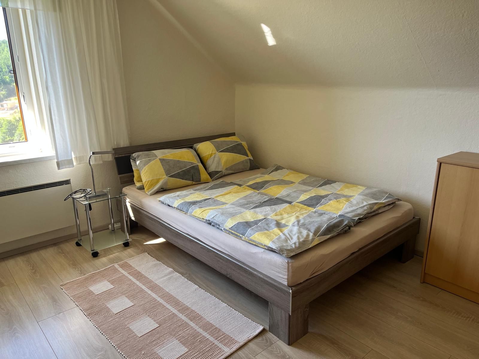 Útulná spálňa s manželskou posteľou, vzorovanými perinami, malým nočným stolíkom a oknom so závesmi.