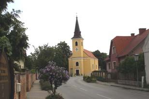 Žltý kostol s vežou v tichej ulici, obklopený stromami a domami.