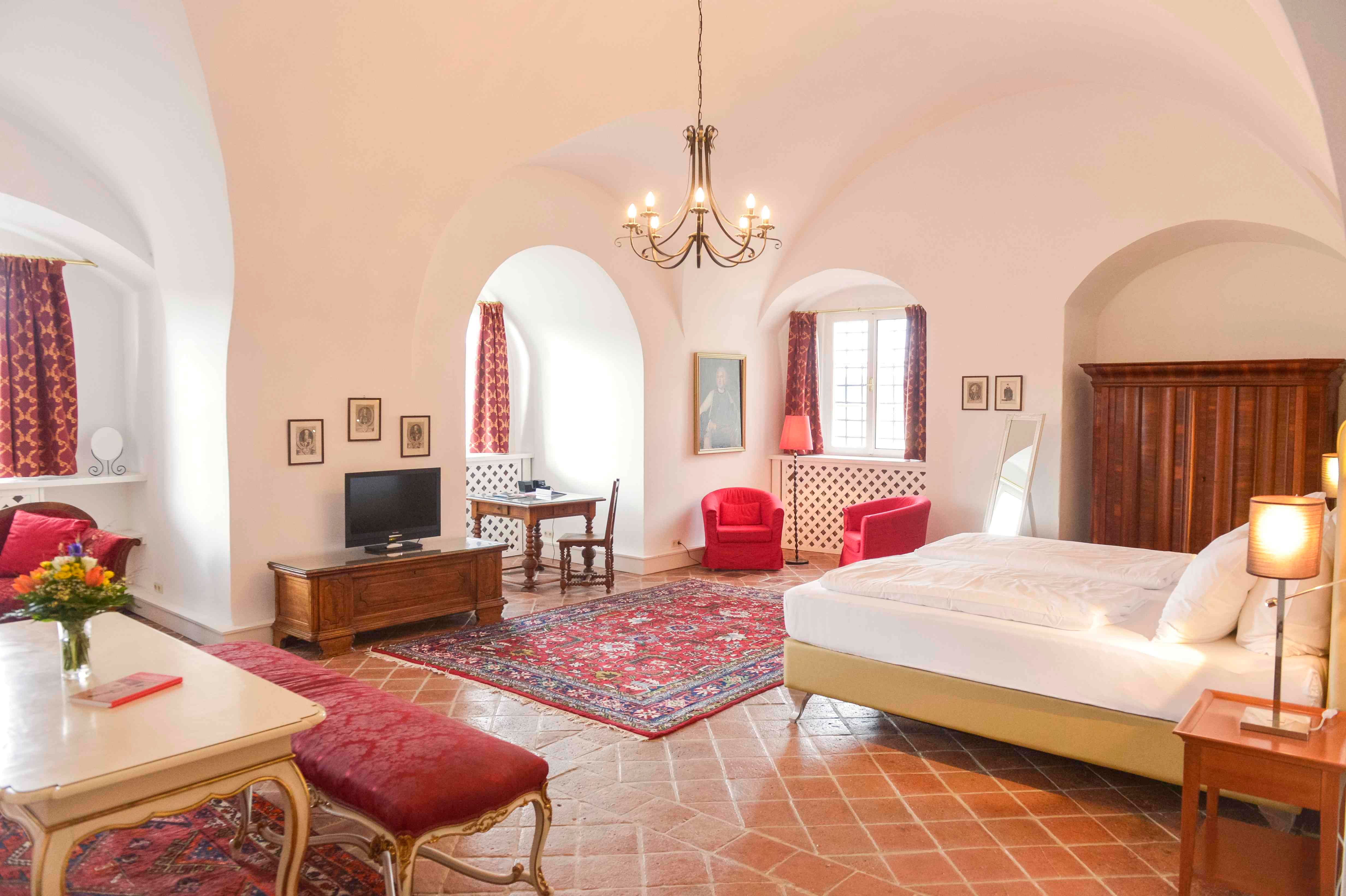 Luxusný apartmán so starožitným nábytkom, červenými kobercami a závesmi, veľkou posteľou a lustrom.