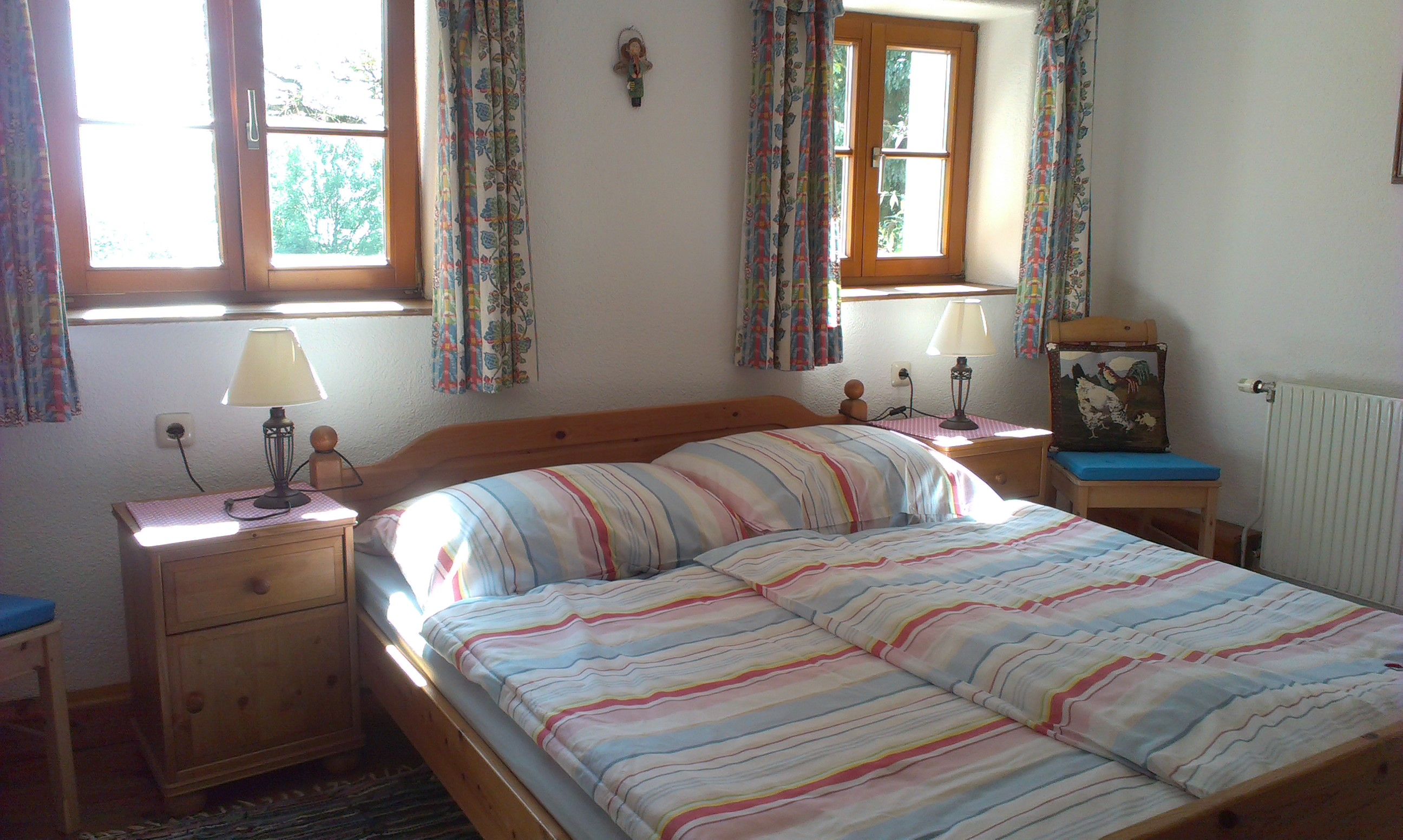 Útulná spálňa s manželskou posteľou, pruhovanou posteľnou bielizňou, dvoma nočnými stolíkmi s lampami a farebnými závesmi na oknách.