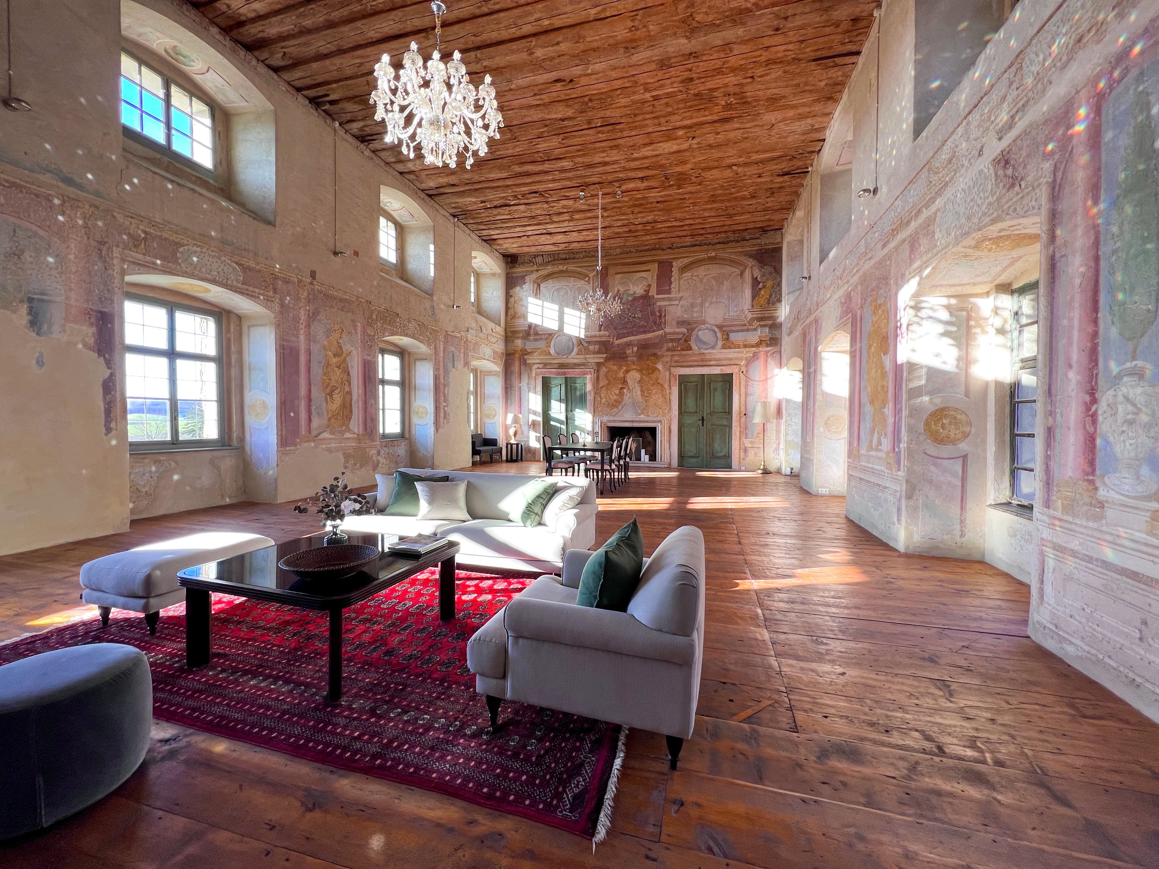 Historická tanečná sála s drevenou podlahou, maľovanými stenami a lustrom. V miestnosti je umiestnený moderný nábytok a červený koberec.