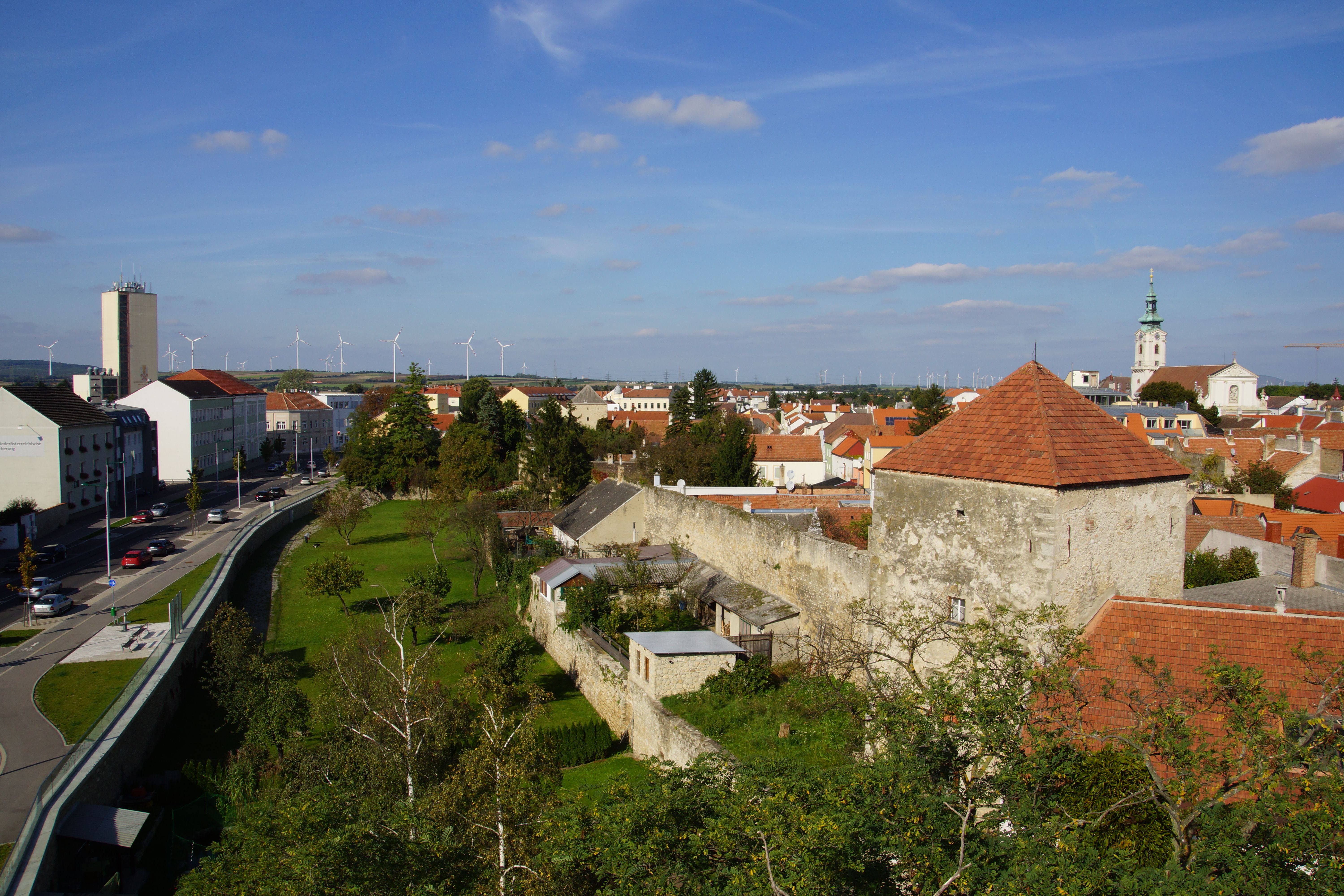Pohľad na mesto Bruck an der Leitha s mestskými hradbami, kostolom a veternými turbínami v pozadí.