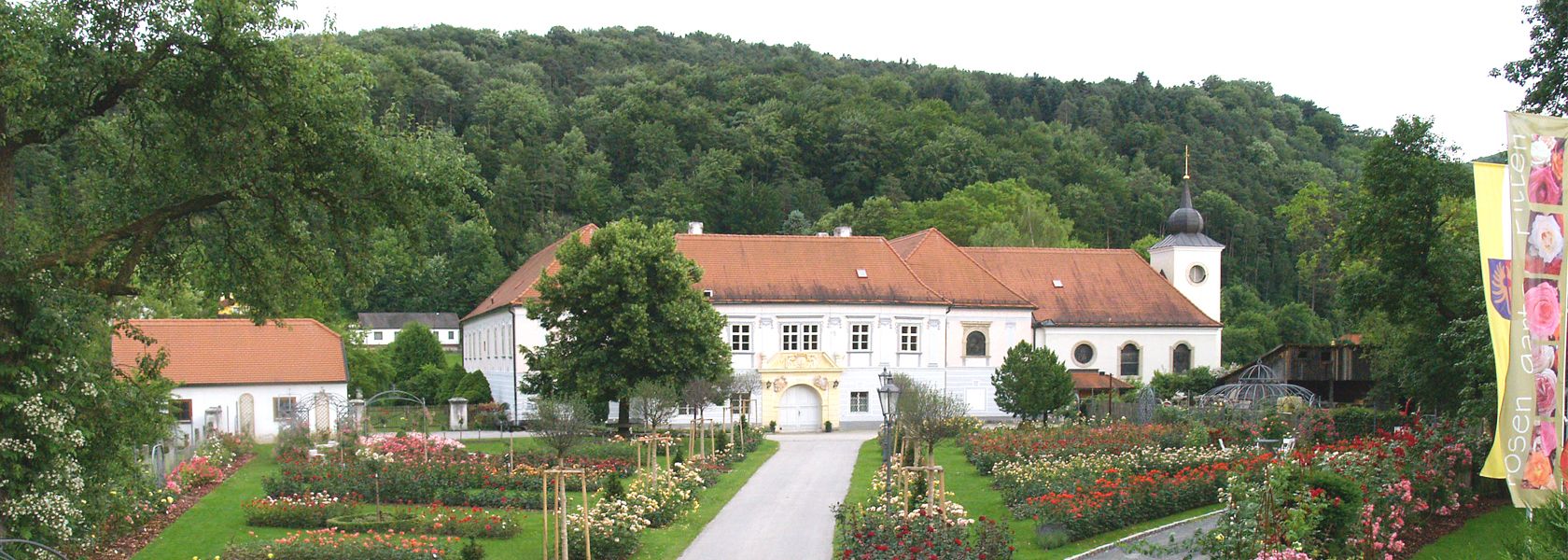 Historická budova s červenou strechou, obklopená ružovou záhradou a stromami, v pozadí zalesnený kopec.