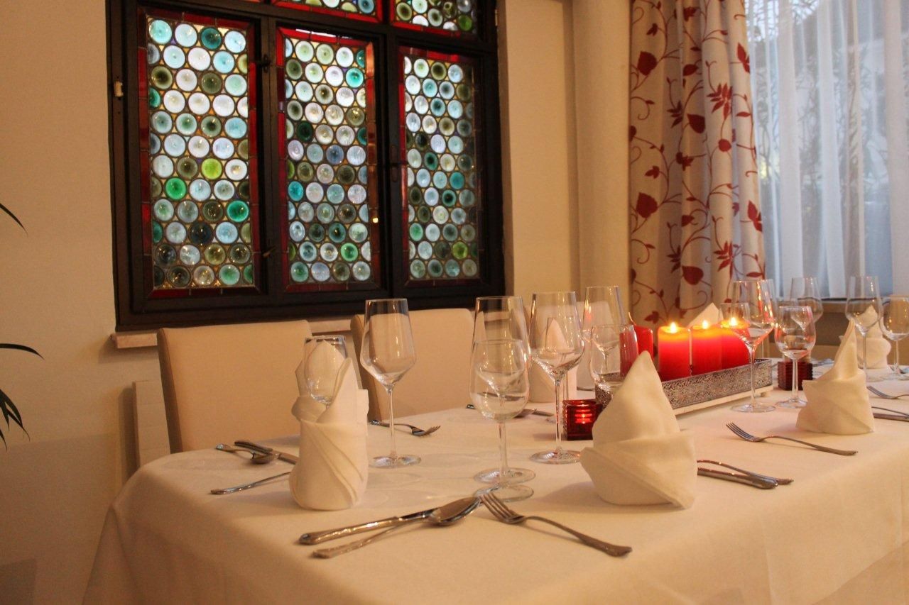 Elegantný stôl s bielymi obrusmi, pohármi na víno a červenými sviečkami pred farebným oknom.