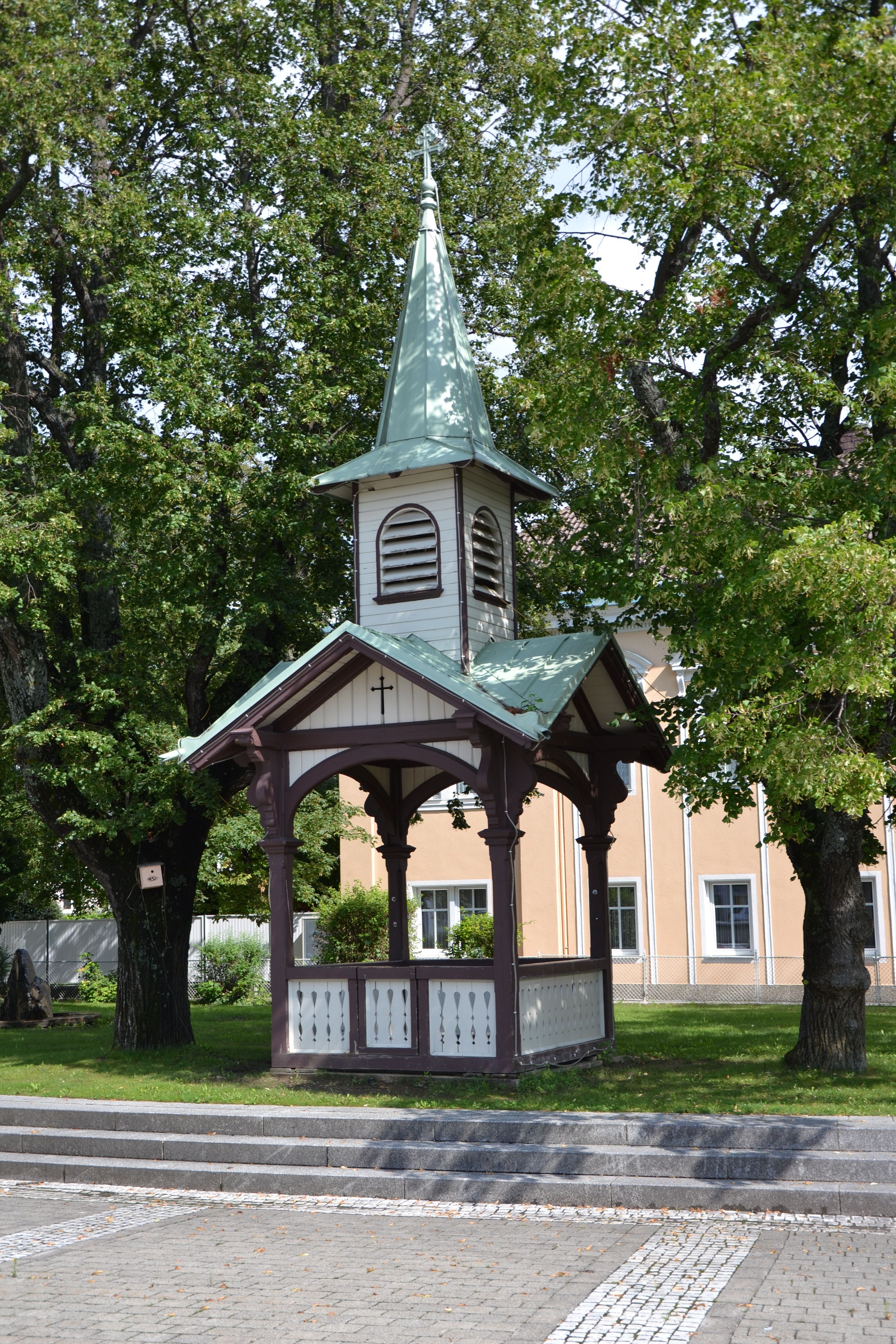 Malý pavilón so špicatou strechou a krížom, obklopený stromami.
